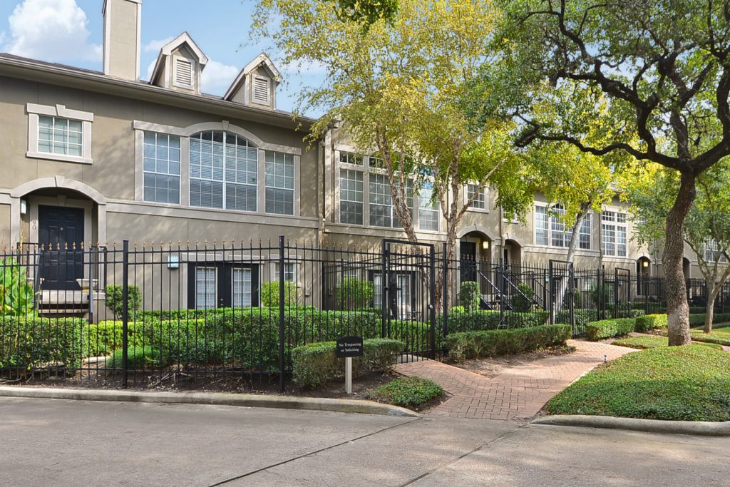 Apartments For Rent Midtown Houston A row of apartment townhouses with large windows and neatly trimmed hedges, partially obscured by a black iron fence, is framed by trees and a paved walkway in the foreground.