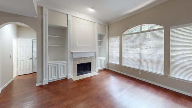 Apartments For Rent Midtown Houston An empty living room features a fireplace flanked by built-in shelves, a large window with blinds, wooden floors, and an arched doorway leading to another room.